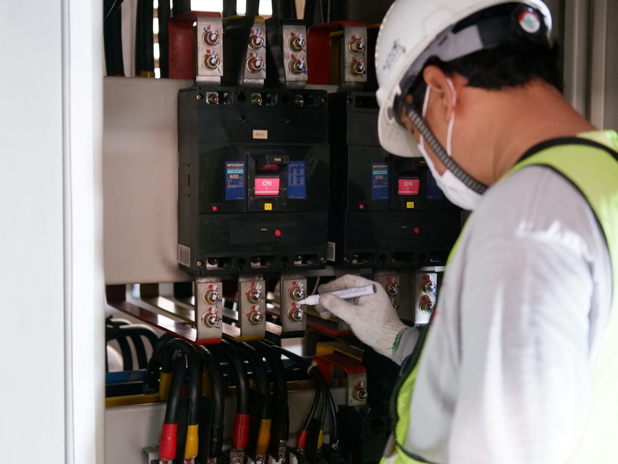 Technician in white helmet recording readings on a clipboard while inspecting an electrical switchboard