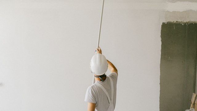 Painter in white overalls and hard hat repainting an interior wall with a roller